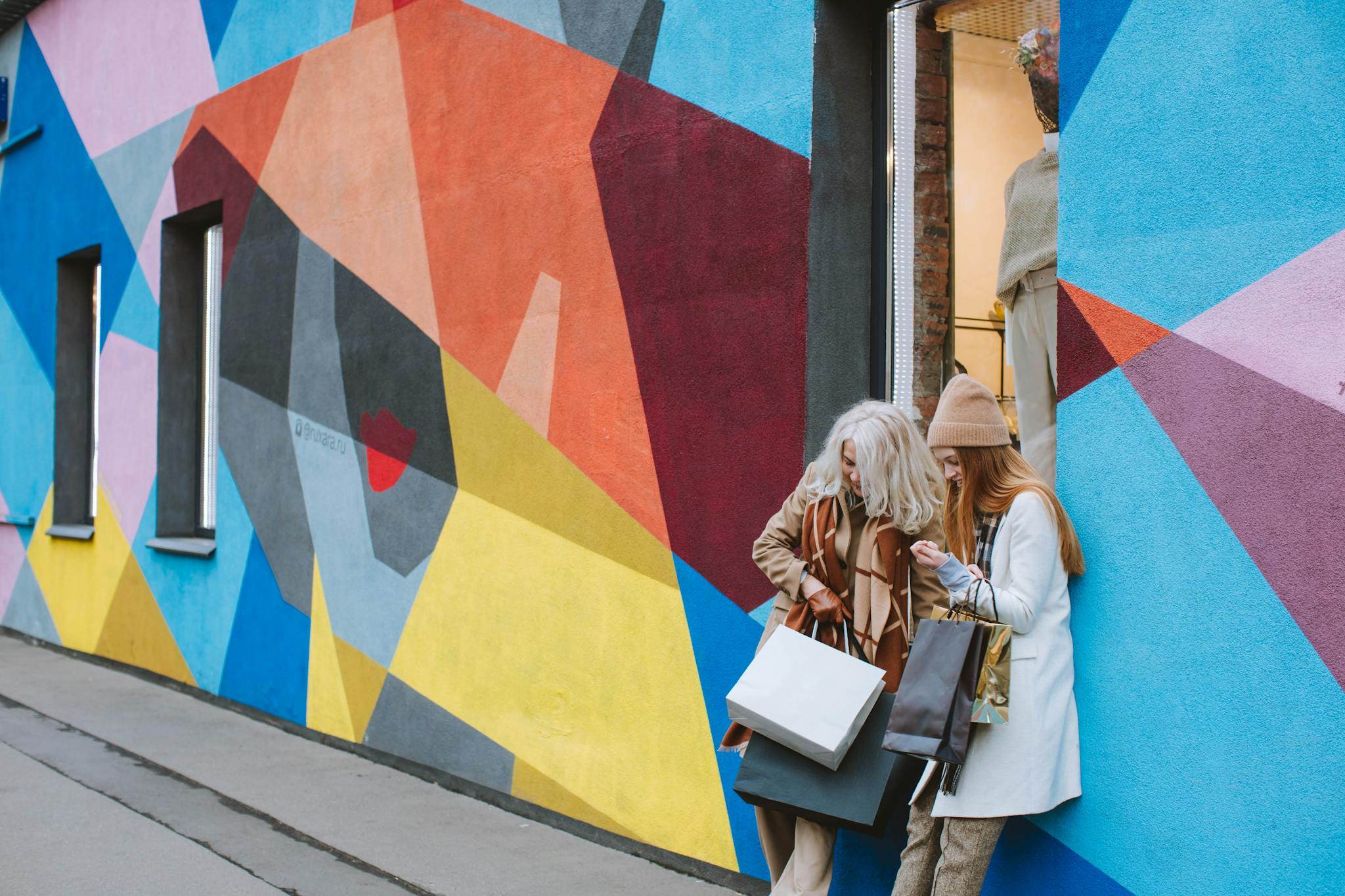 Two women enjoy a shopping day, standing against a vibrant mural with shopping bags.